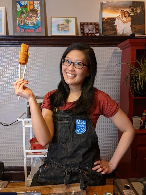 Chef Joyce wearing apron and smiling at camera while holding a spring roll using chopsticks