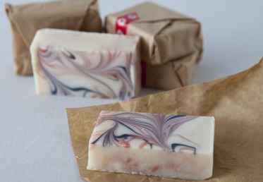 A block of handmade soap sits on a parchment paper wrapper, with more soap packaged in the background